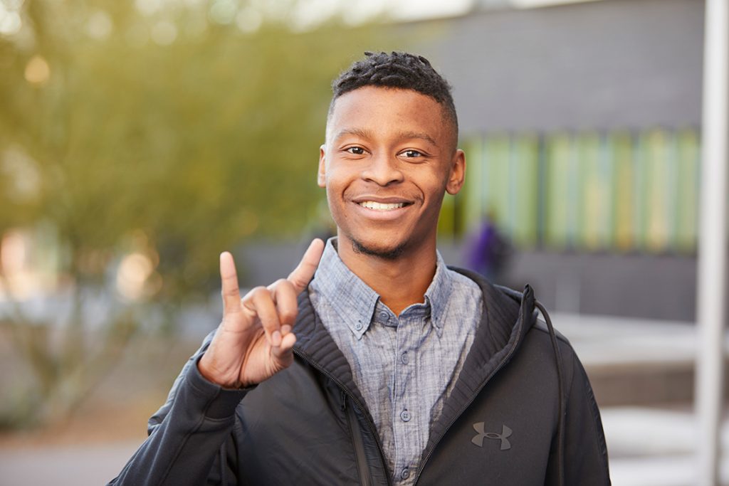 Young man smiling outside giving a lopes up gesture with his hand.