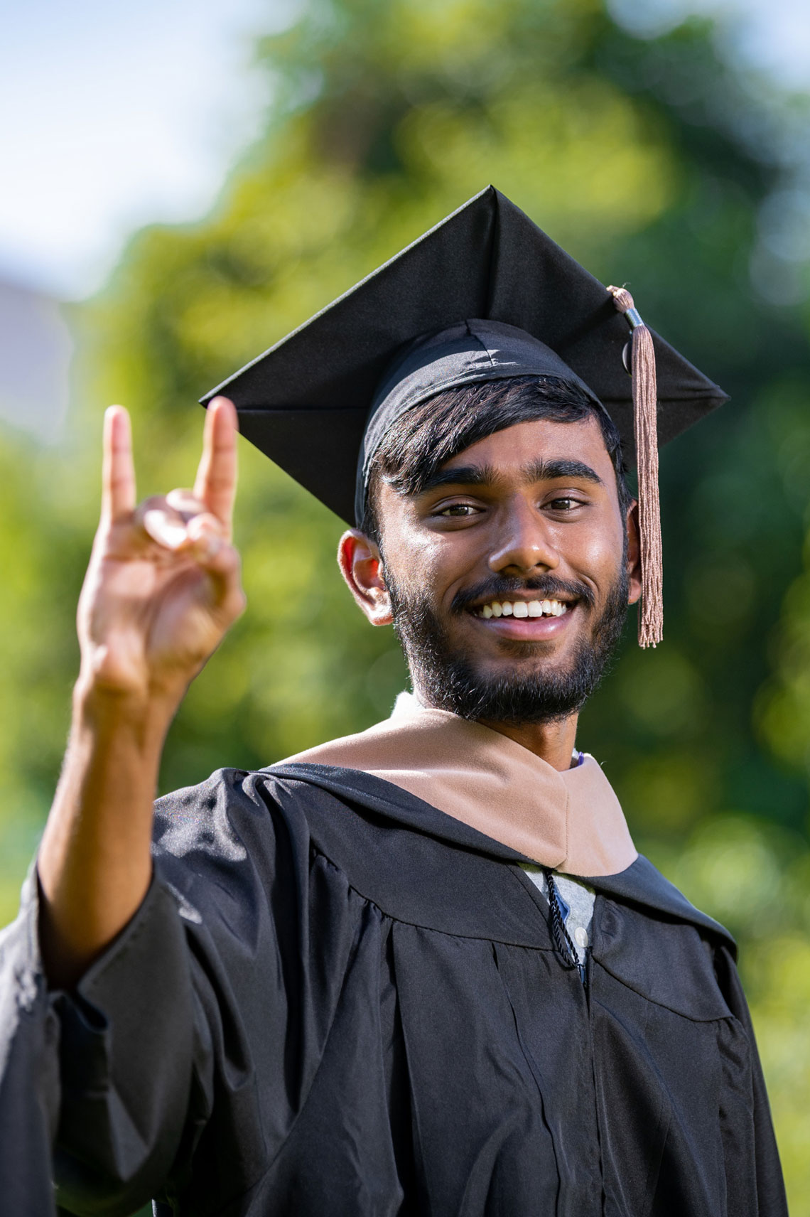 Young man outside in graduation garb giving lopes up gesture and smiling.