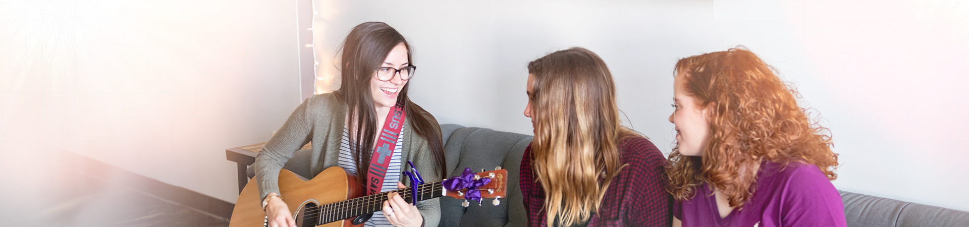 Three young women sitting on couch with guitar.