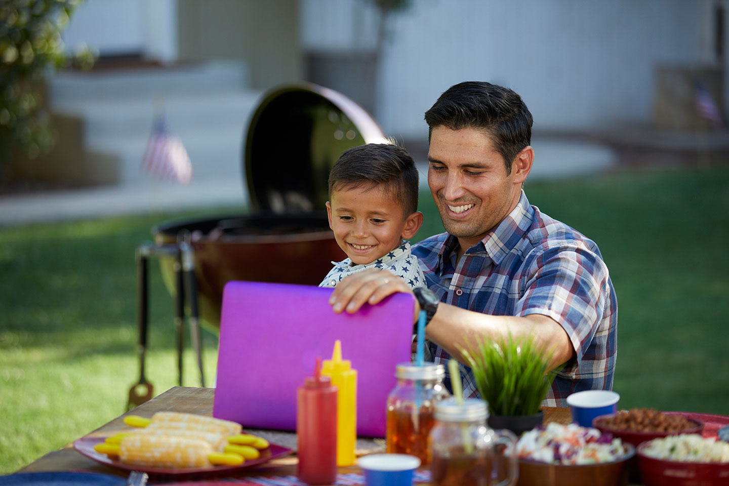 Father and son smiling while working on laptop outside.
