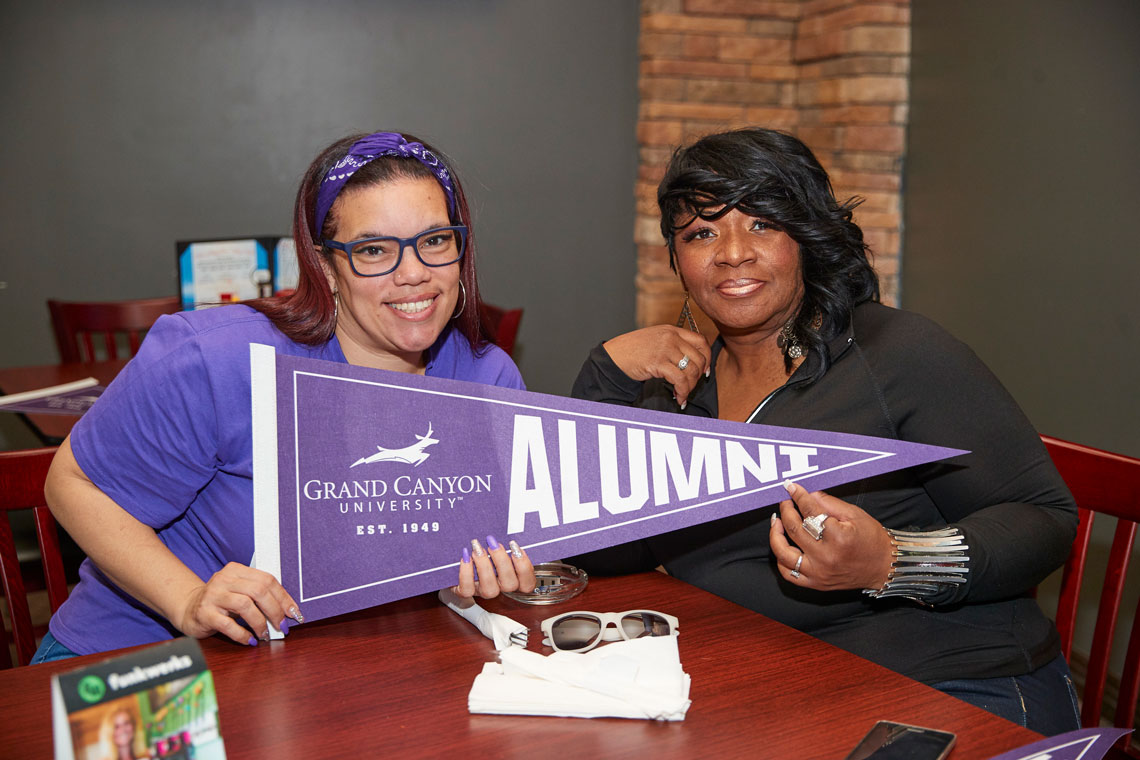 Two women sitting at desk holding GCU Alumni flag while smiling.